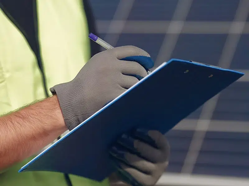 Personne avec gants écrivant sur un clipboard devant panneau solaire
